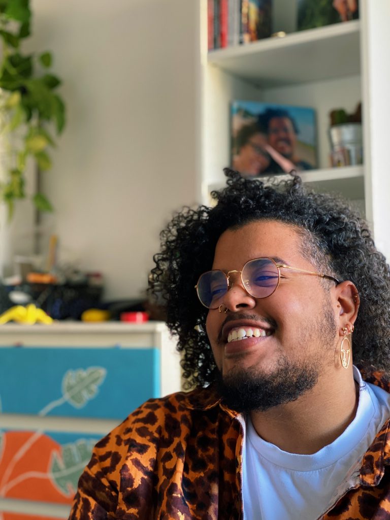 The author of the post as an adult. They have curly afro hair and a beard. They are smiling broadly and looking off to the side. They are wearing round glasses and a white t-shirt underneath a shirt with an orange and brown tiger print. 