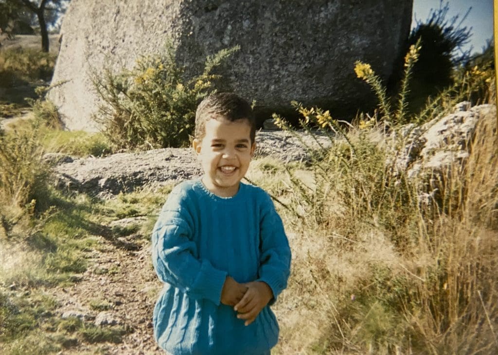 A young child wearing a blue knitted sweater smiles at the camera. They have brown hair, brown eyes and brown skin and are standing in front of green shrubs and rocks