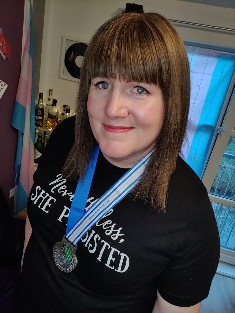 Danielle, a woman with long brown hair, wears her medal on top of a t-shirt which reads 'nevertheless, she persisted'