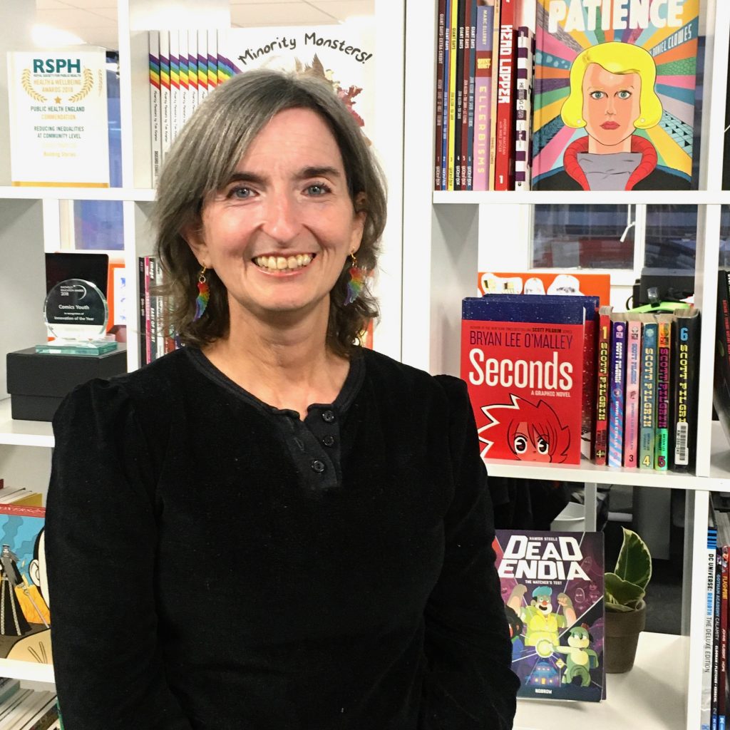 A white woman with grey hair and colourful earrings stands in front of a bookshelf filled with colourful books