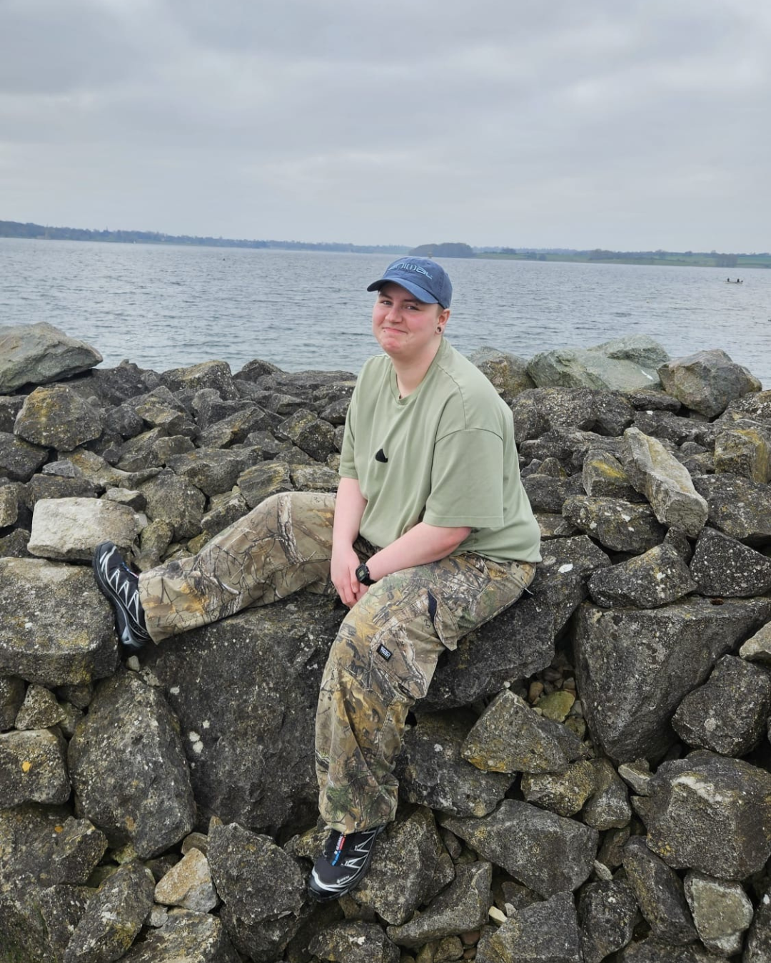 Newton sits on some rocks next to the sea on an overcast day. He's wearing a blue cap, a pale green tee and camo trousers.