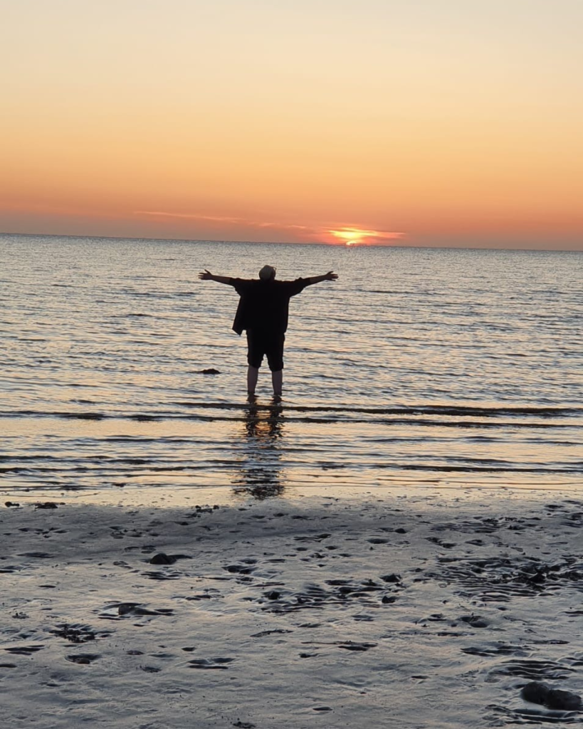 Newton stands ankle-deep in the sea on a beach at sunset, arms outstretched.