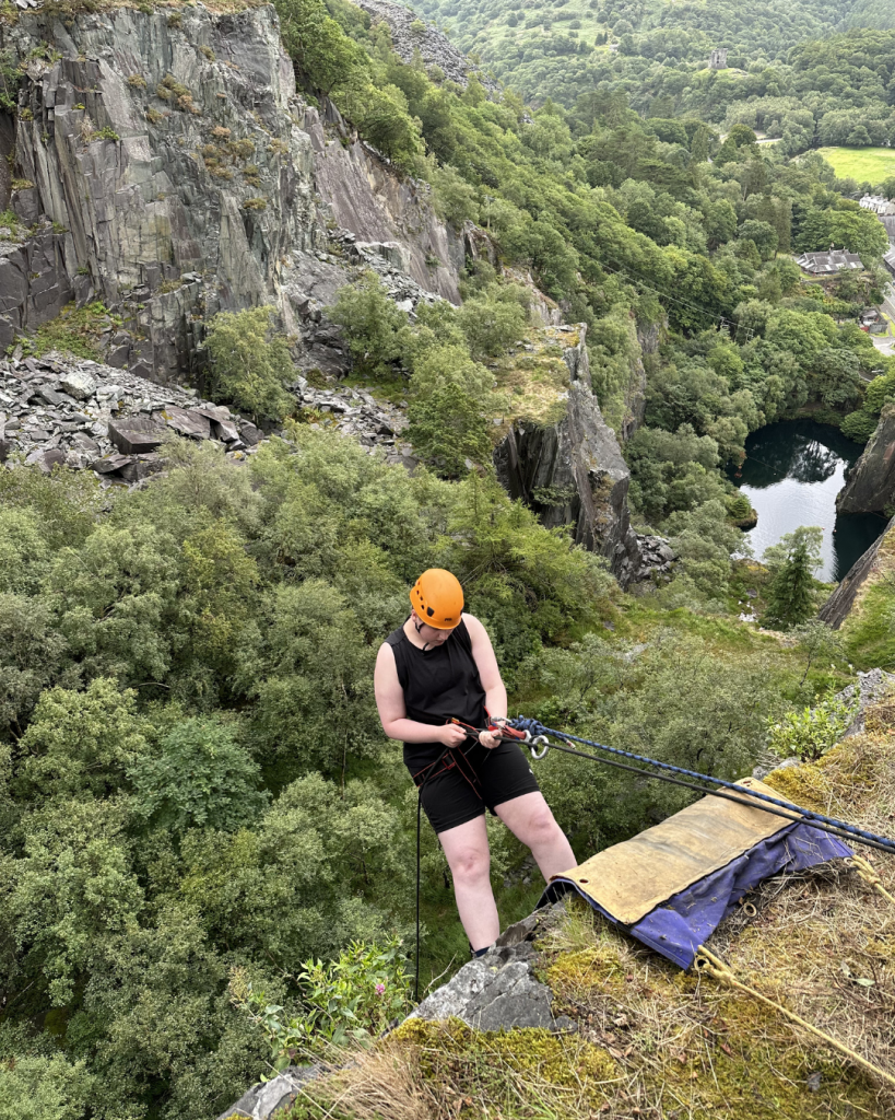 Newton abseils down a cliff, wearing a black tank top and shorts and an orange helmet. Behind him is lush green forest and more cliffs.