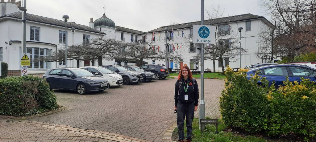 Eli stands smiling next to a sign that says 'Pax Lodge' under the Girlguiding logo. In the background is a car park and a white, three storey building.