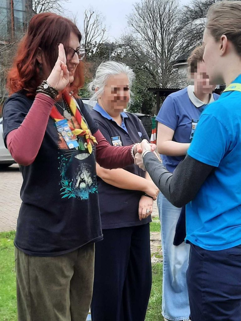 Eli raises one hand, palm outward, with their three middle fingers up, in the Girlguiding salute.