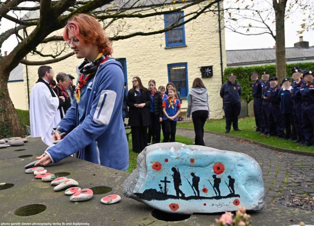 Eli places a stone decorated with a red poppy design next to a larger stone that's painted with a scene of soldiers marching surrounded by poppies, at a World War II remembrance event. In the background, other Guides and Cadets stand.